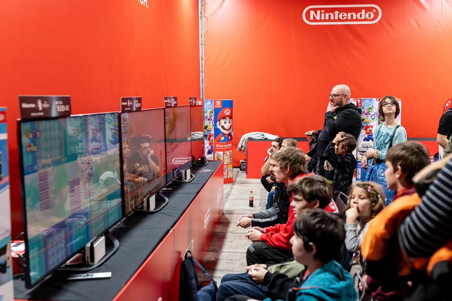People watching video screens at a Nintendo event booth with a large red Nintendo branded wall.