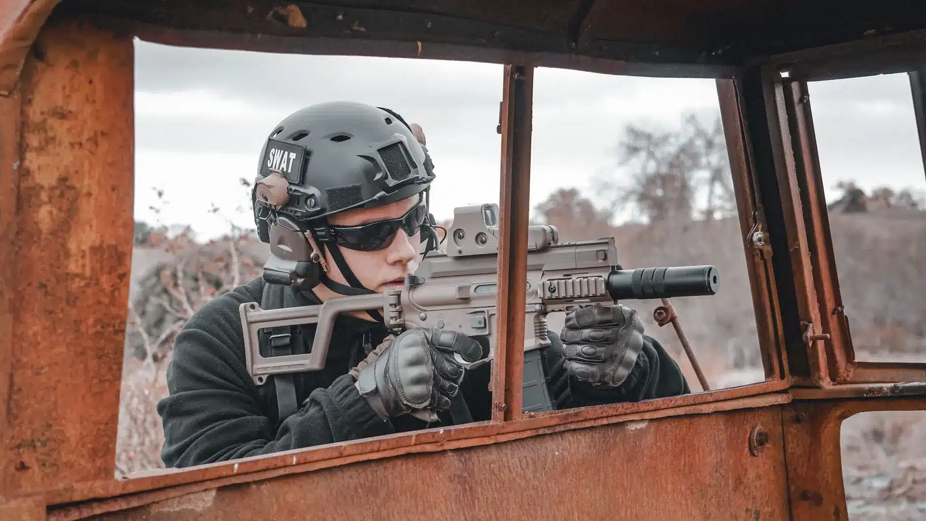 Person in SWAT tactical gear and helmet aiming a suppressed submachine gun through a rusty vehicle window